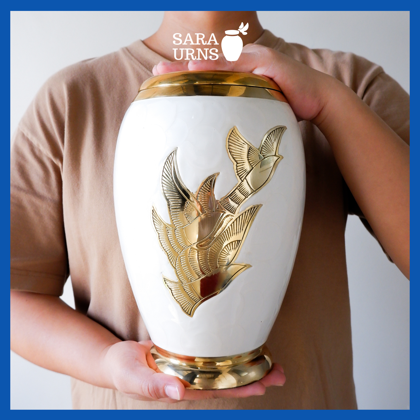 A presentation of a white polished brass metal urn with golden, hand-etched winged doves held by an adult woman with two hands.