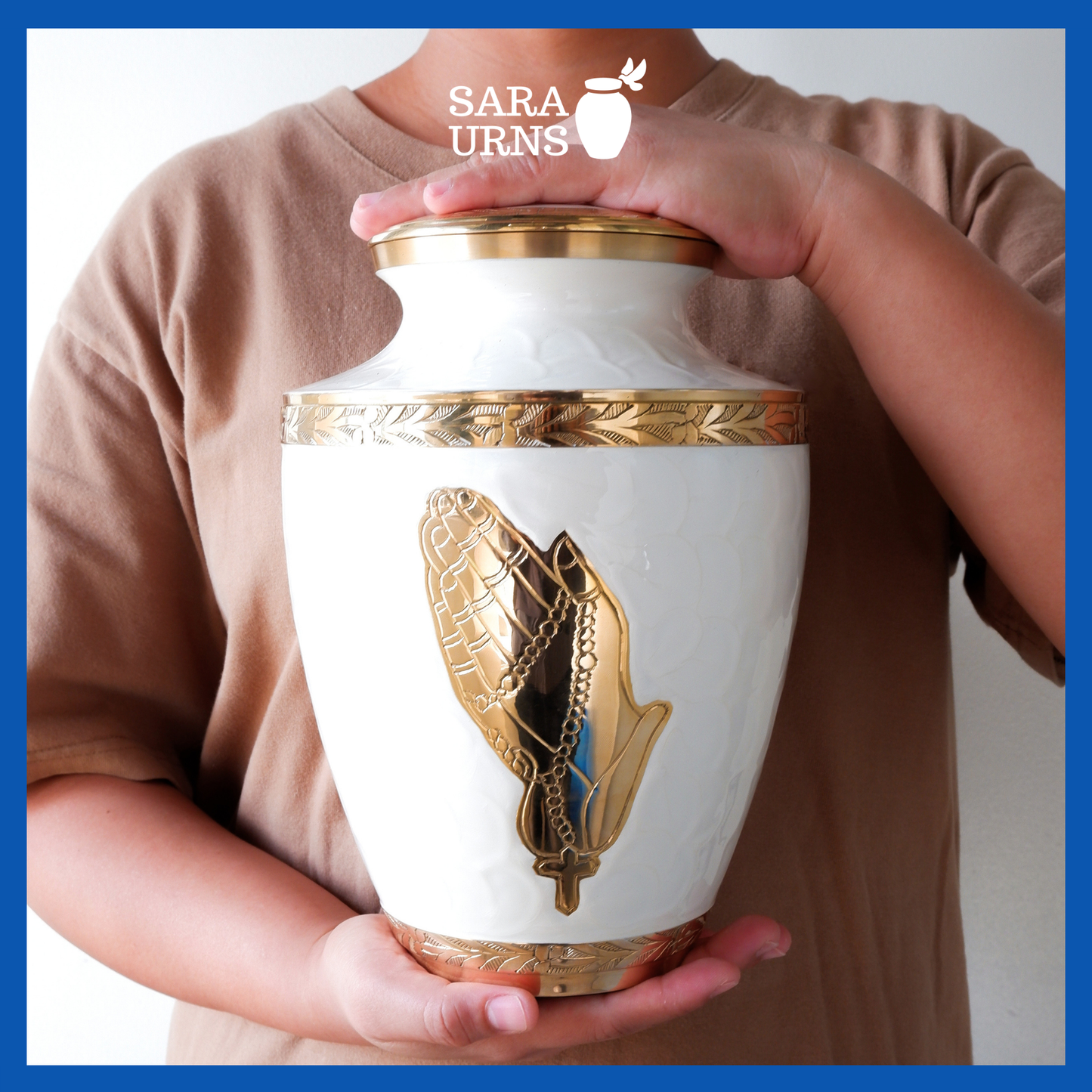 A presentation of a white polished brass metal urn with hand-etched golden praying hands and details held by an adult woman with two hands.