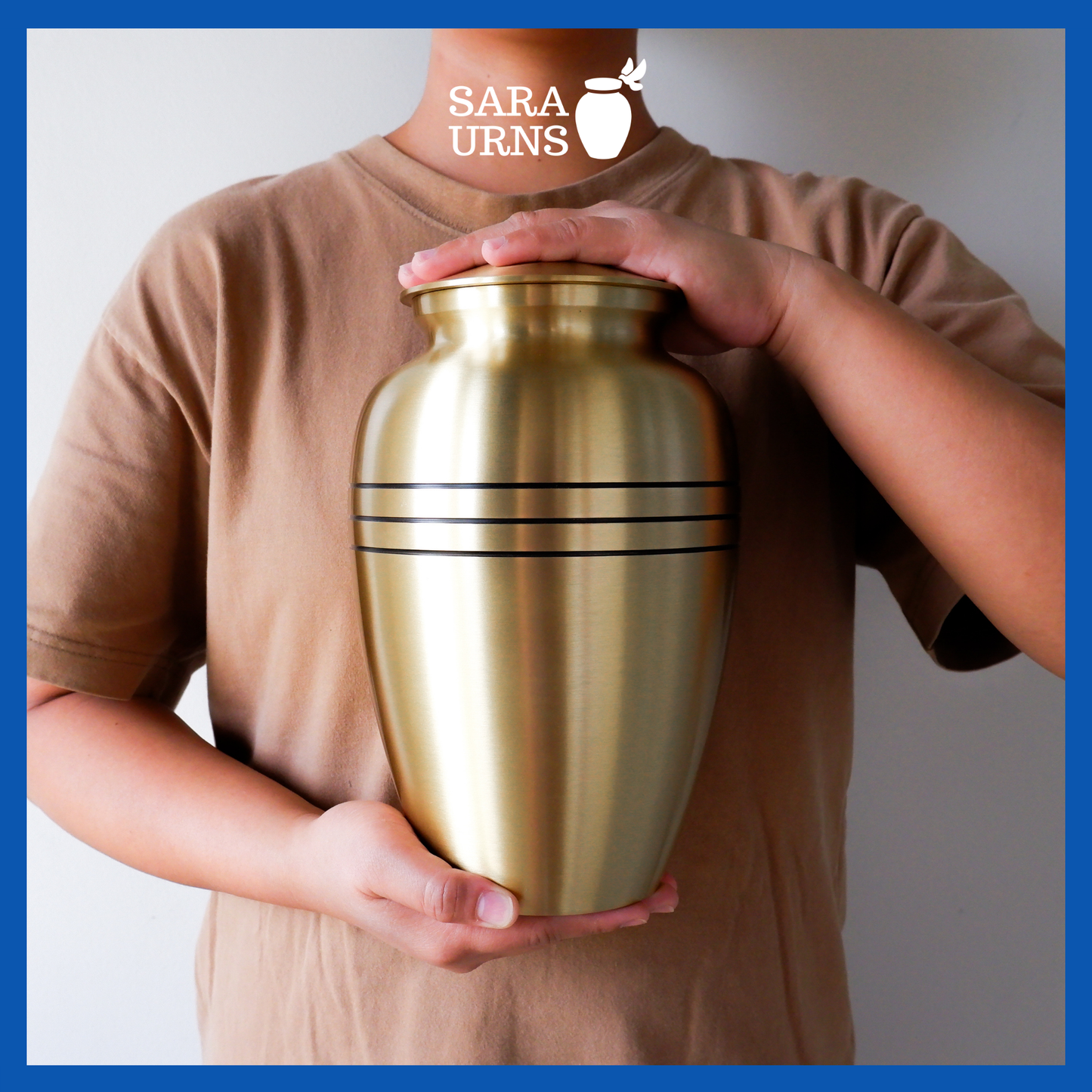A presentation of a gold polished brass metal urn with three, thin black stripes held by an adult woman with two hands.