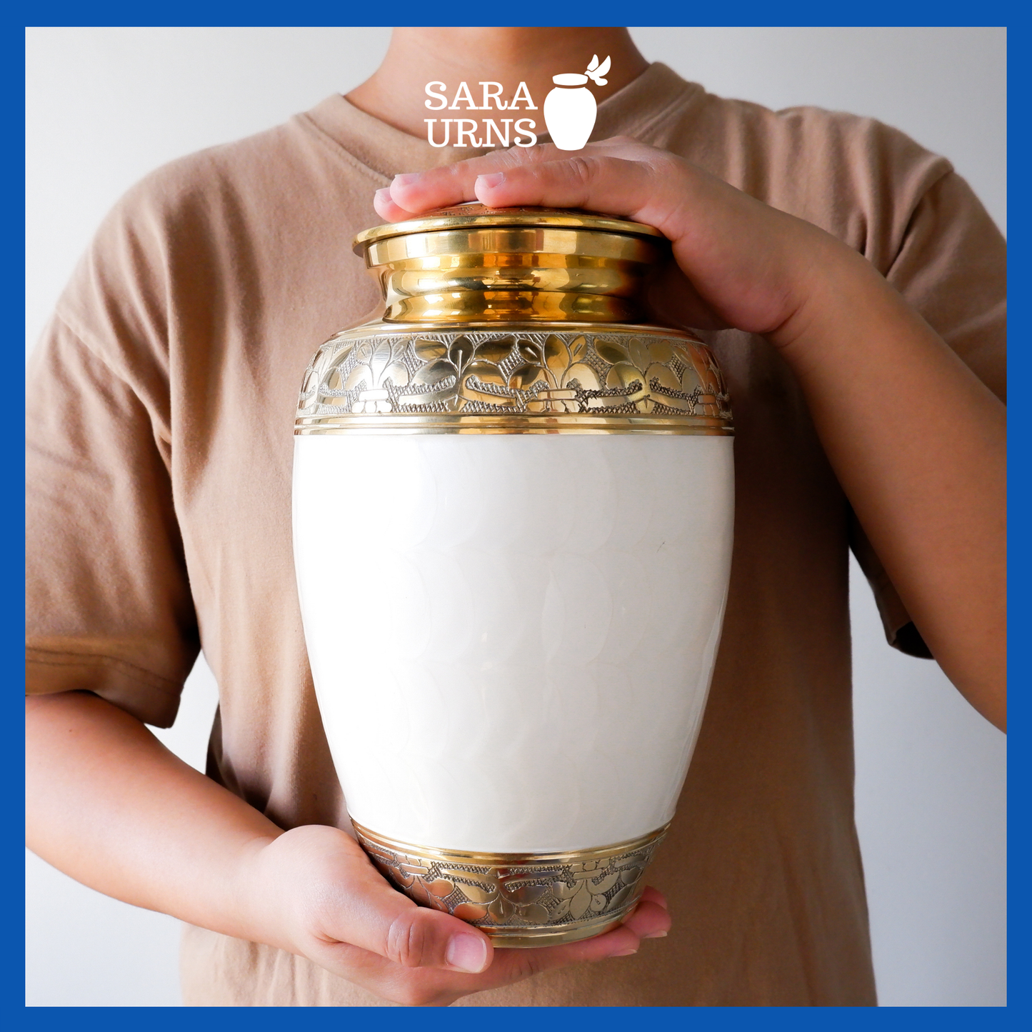 A presentation of a white polished brass metal urn with a golden, hand-etched leaf pattern detail held by an adult woman with two hands.