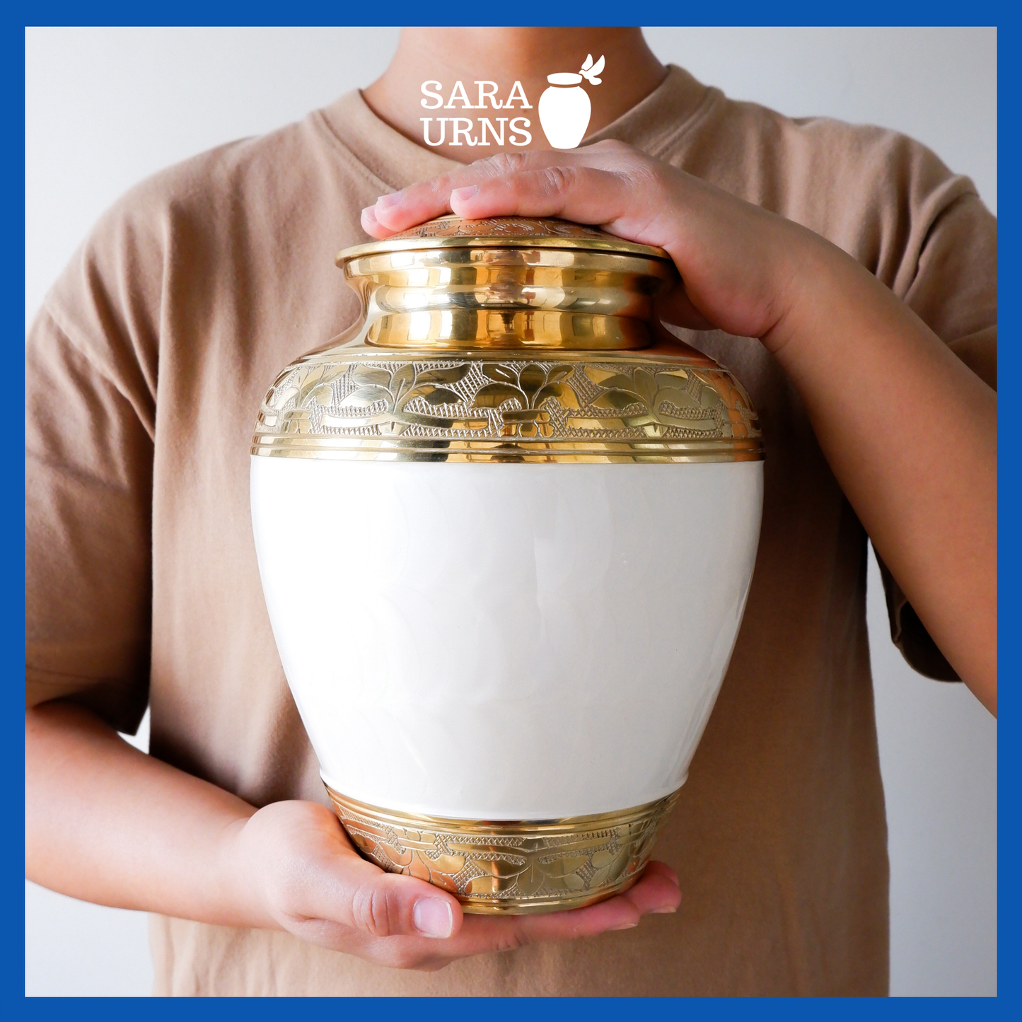A presentation of a white polished brass metal urn with a golden, hand-etched leaf pattern detail held by an adult woman with two hands.