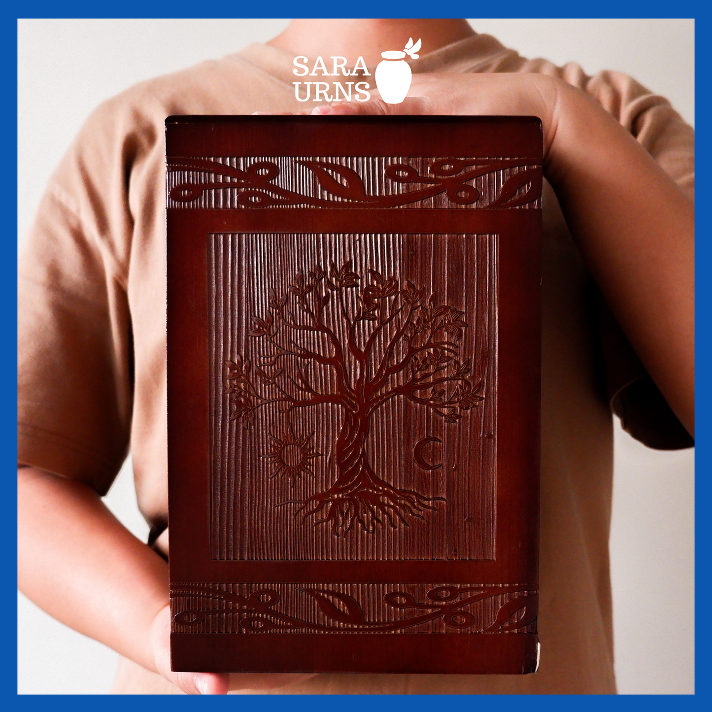 A presentation of a brown pine wooden urn with an intricate engraving of the tree of life held by an adult woman with two hands.