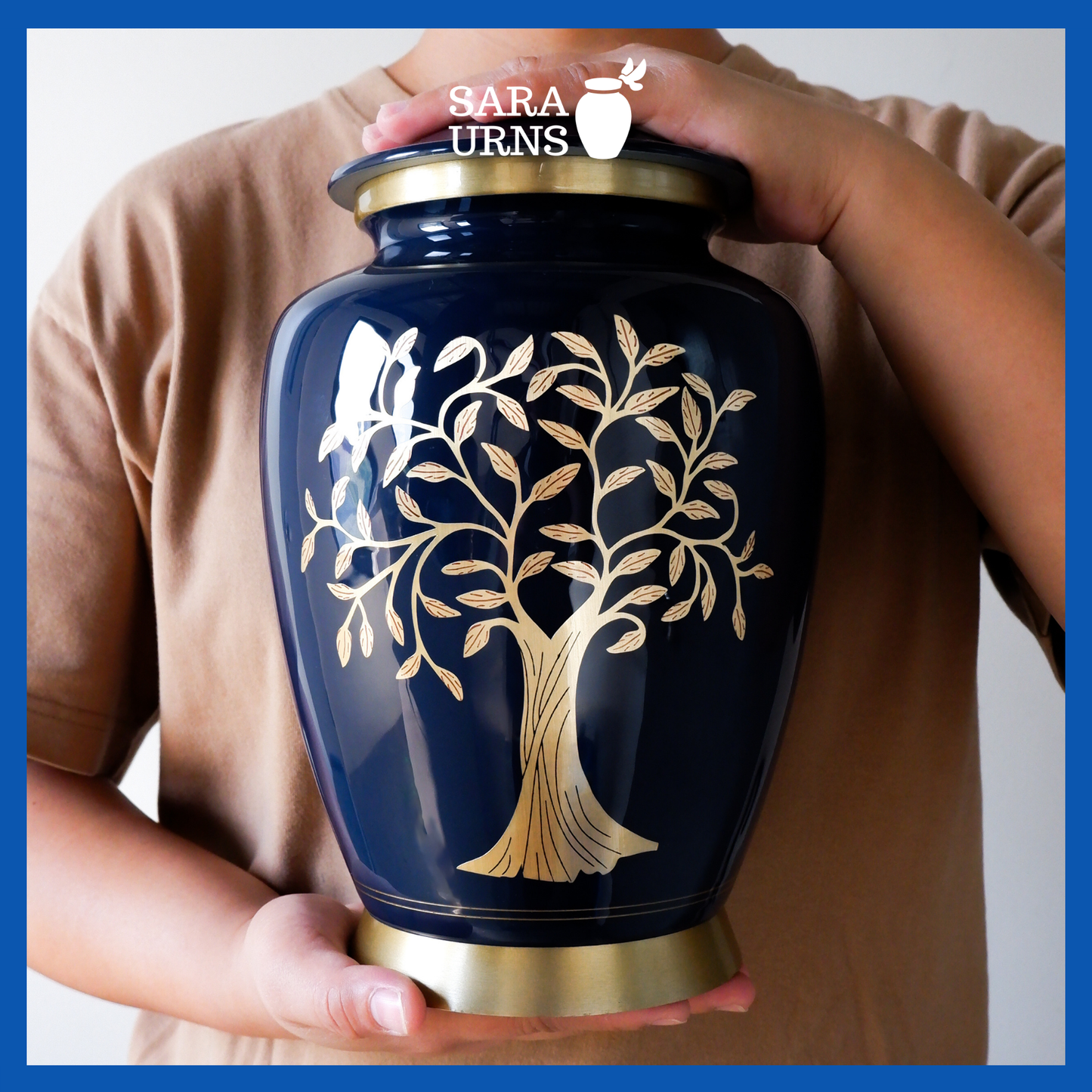 A presentation of a blue polished aluminum metal urn with a golden, hand-etched tree of life detail held by an adult woman with two hands.