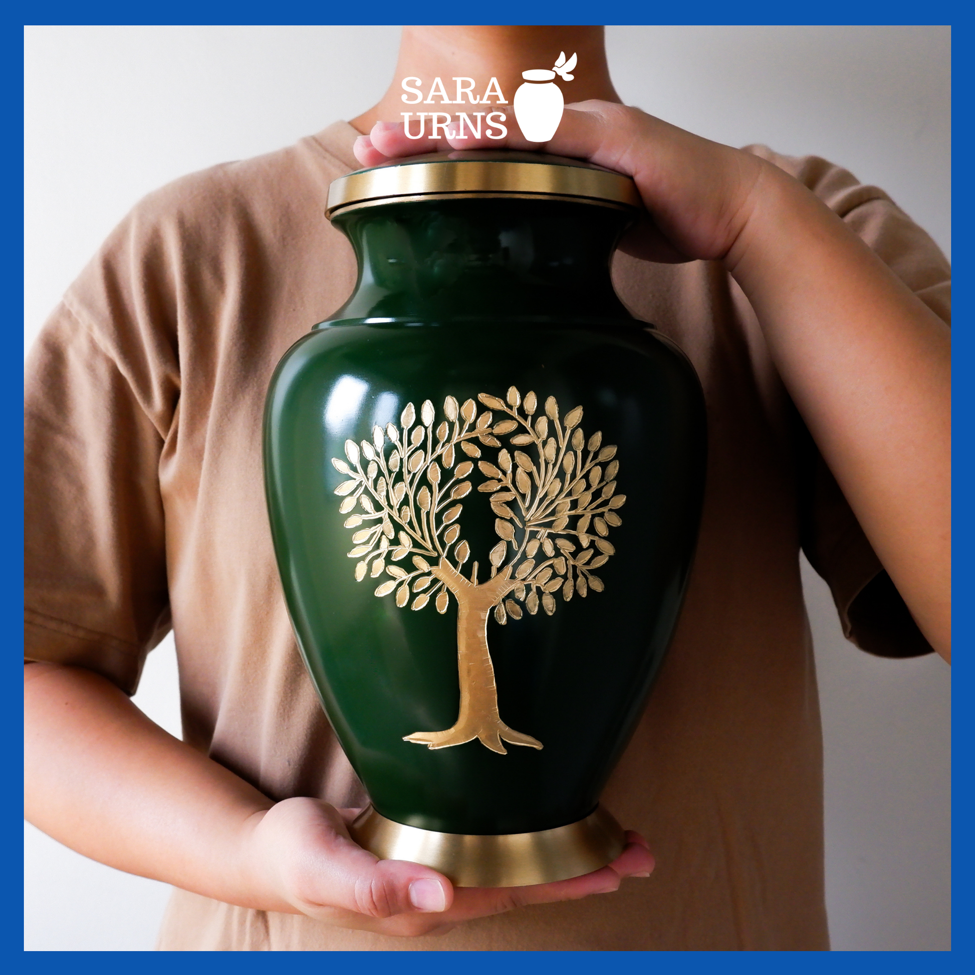 A presentation of a green polished brass metal urn with a golden, hand-etched tree of life detail held by an adult woman with two hands.