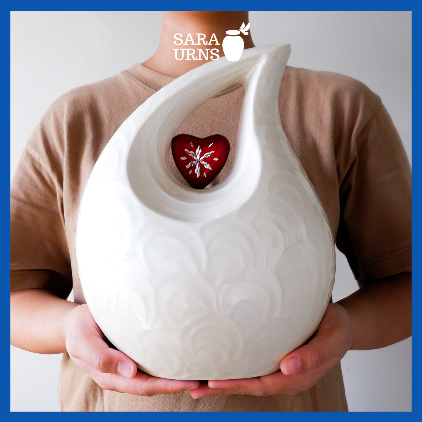 A white polished aluminum metal urn with a droplet shape and heart centerpiece held by an adult woman with two hands.