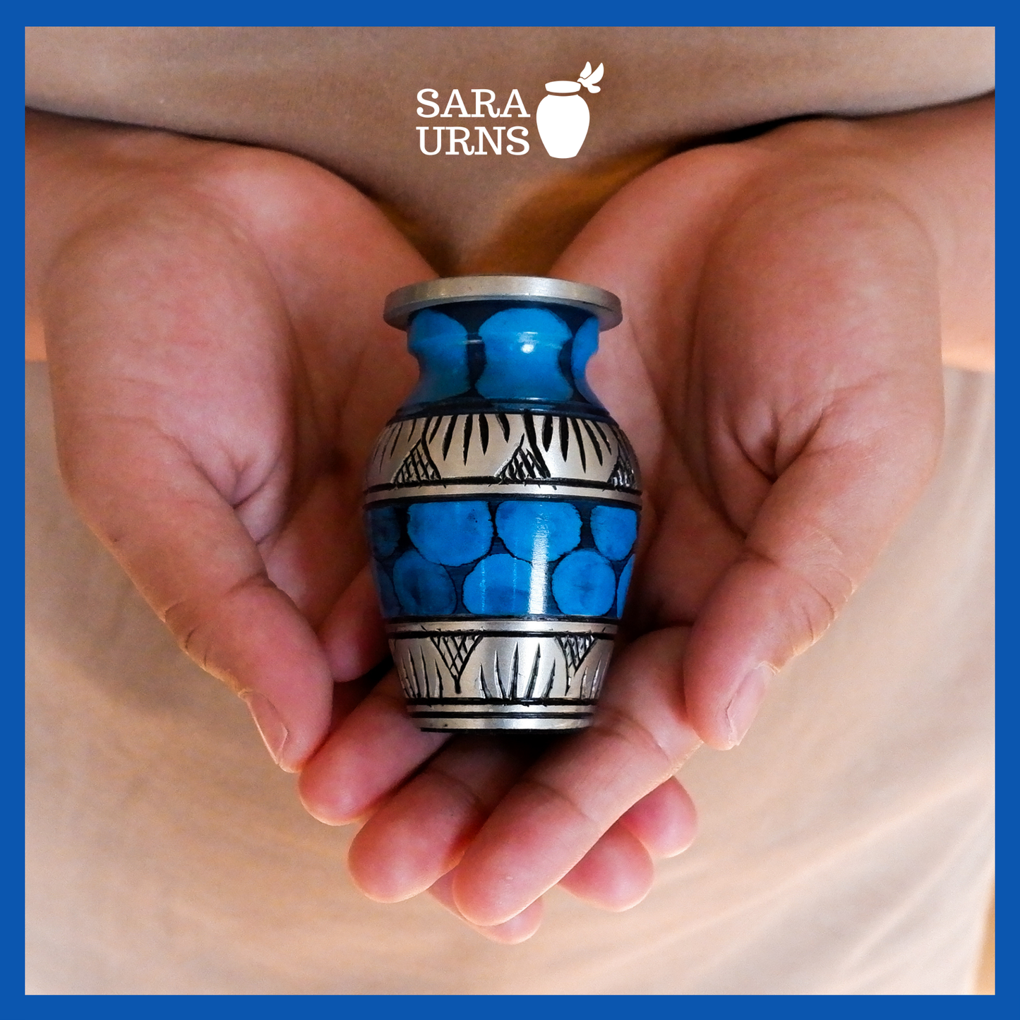 A presentation of a blue aluminum metal keepsake urn with silver hand-etched designs and a painted pattern across the body and lid held by an adult woman with two hands.