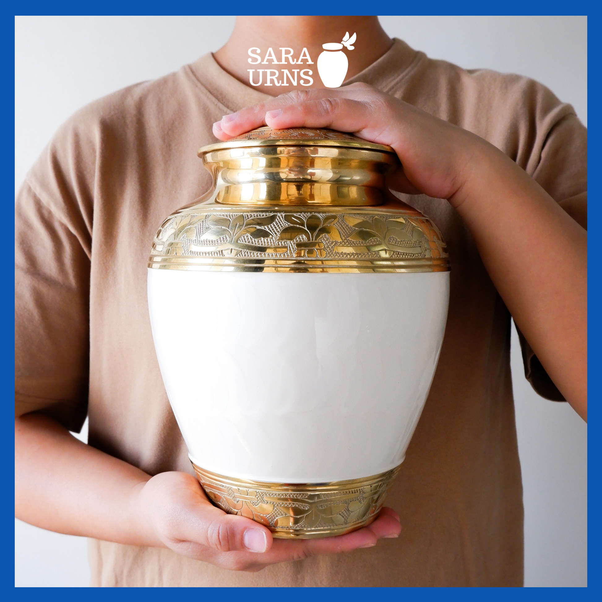 A presentation of a white polished brass metal urn with a golden, hand-etched leaf pattern detail held by an adult woman with two hands.