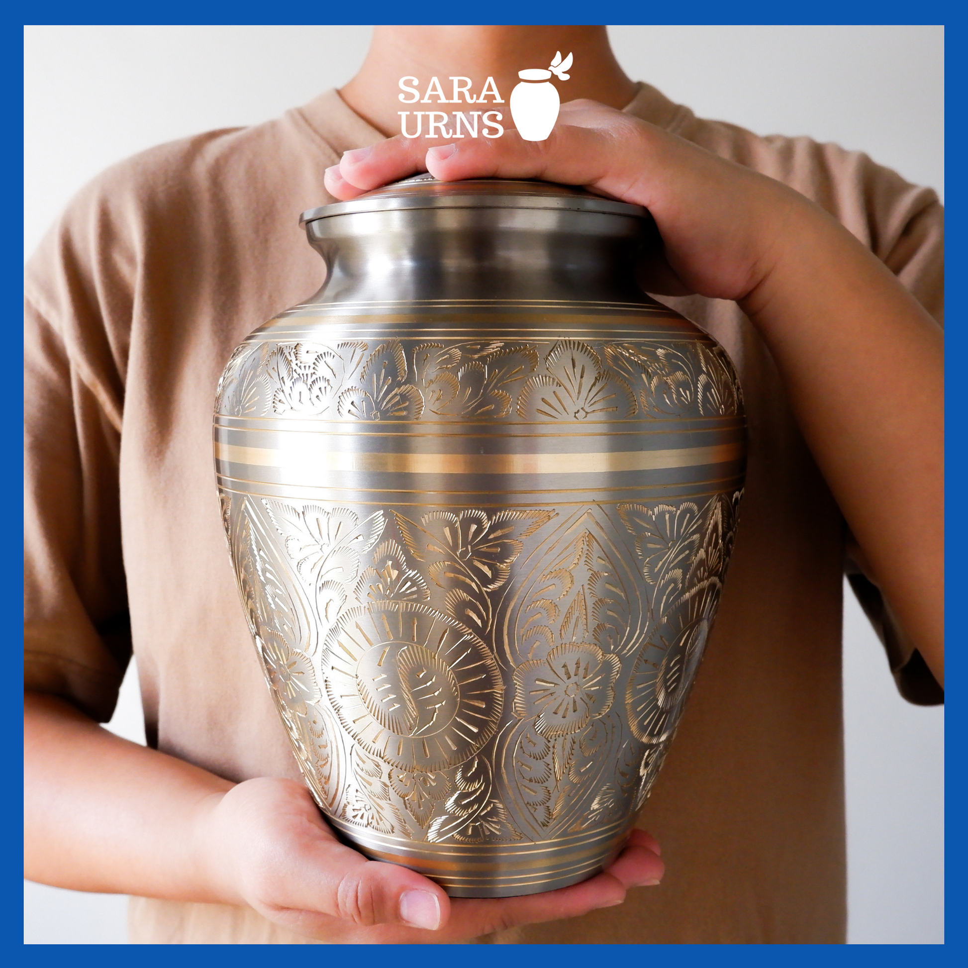 A presentation of a platinum-bronze polished brass metal urn with hand-etched details across the lid and body held by an adult woman with two hands.