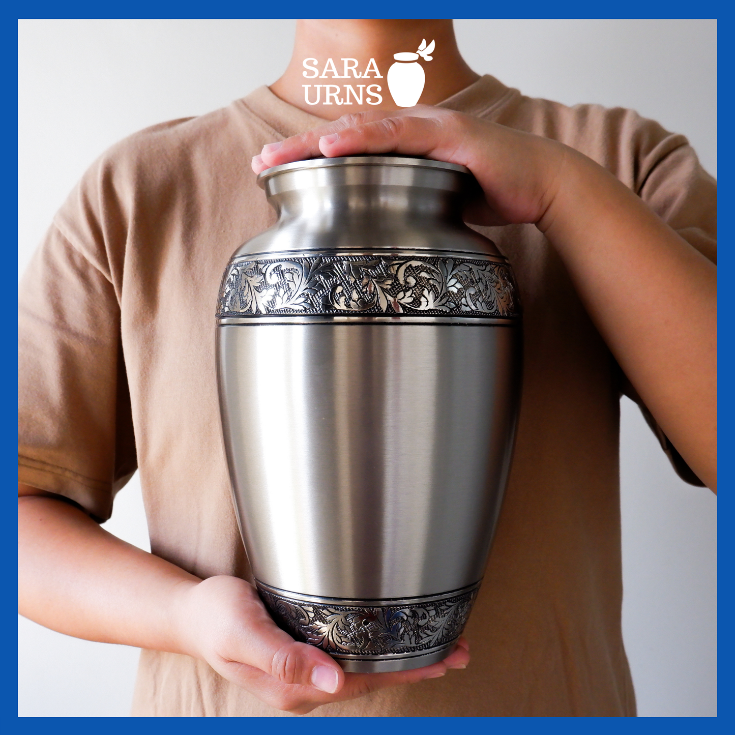 A silver polished brass metal urn with hand-etched details on its body and lid held by an adult woman with two hands.