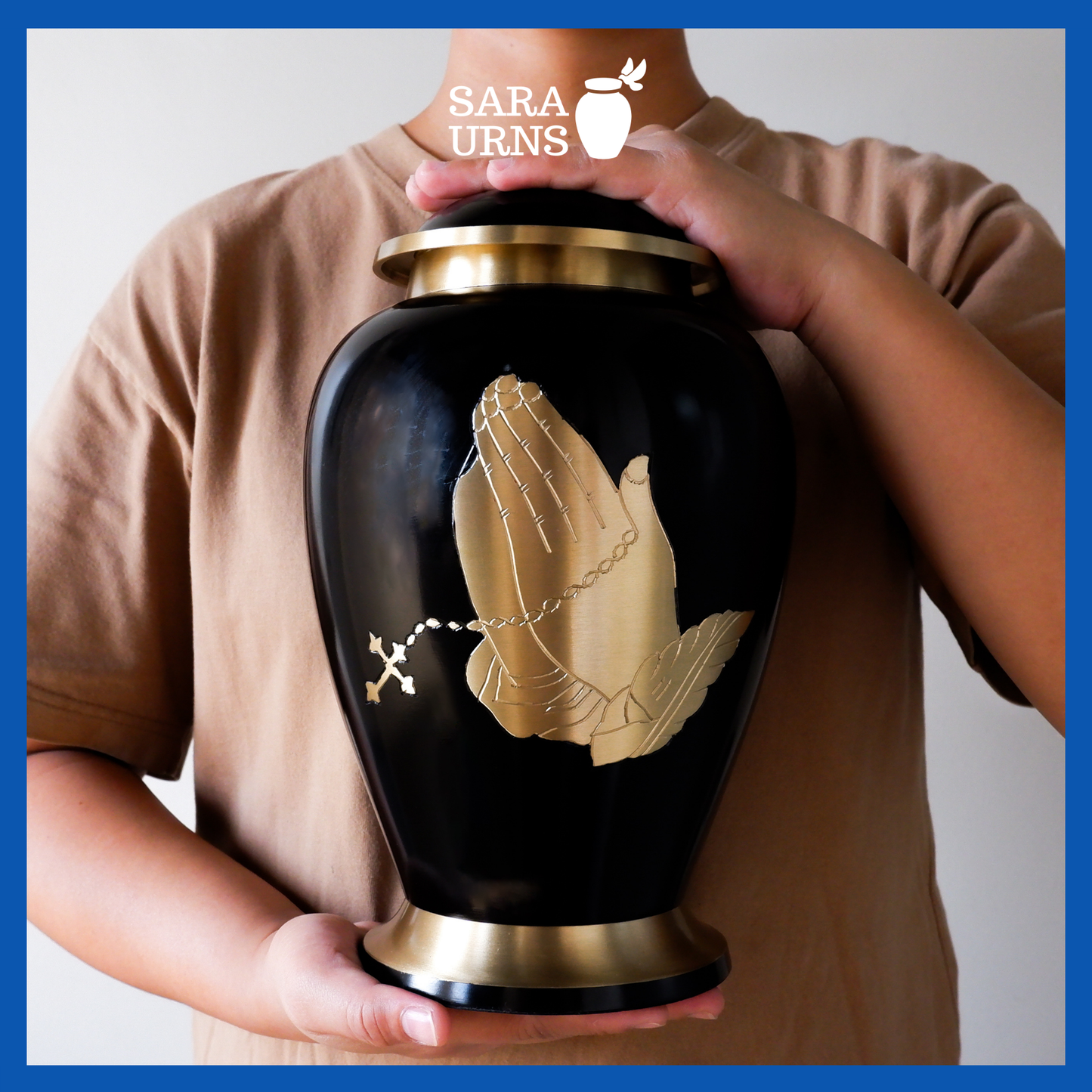 A presentation of a black polished brass metal urn with a golden, hand-etched detail of praying with a rosary held by an adult woman with two hands.