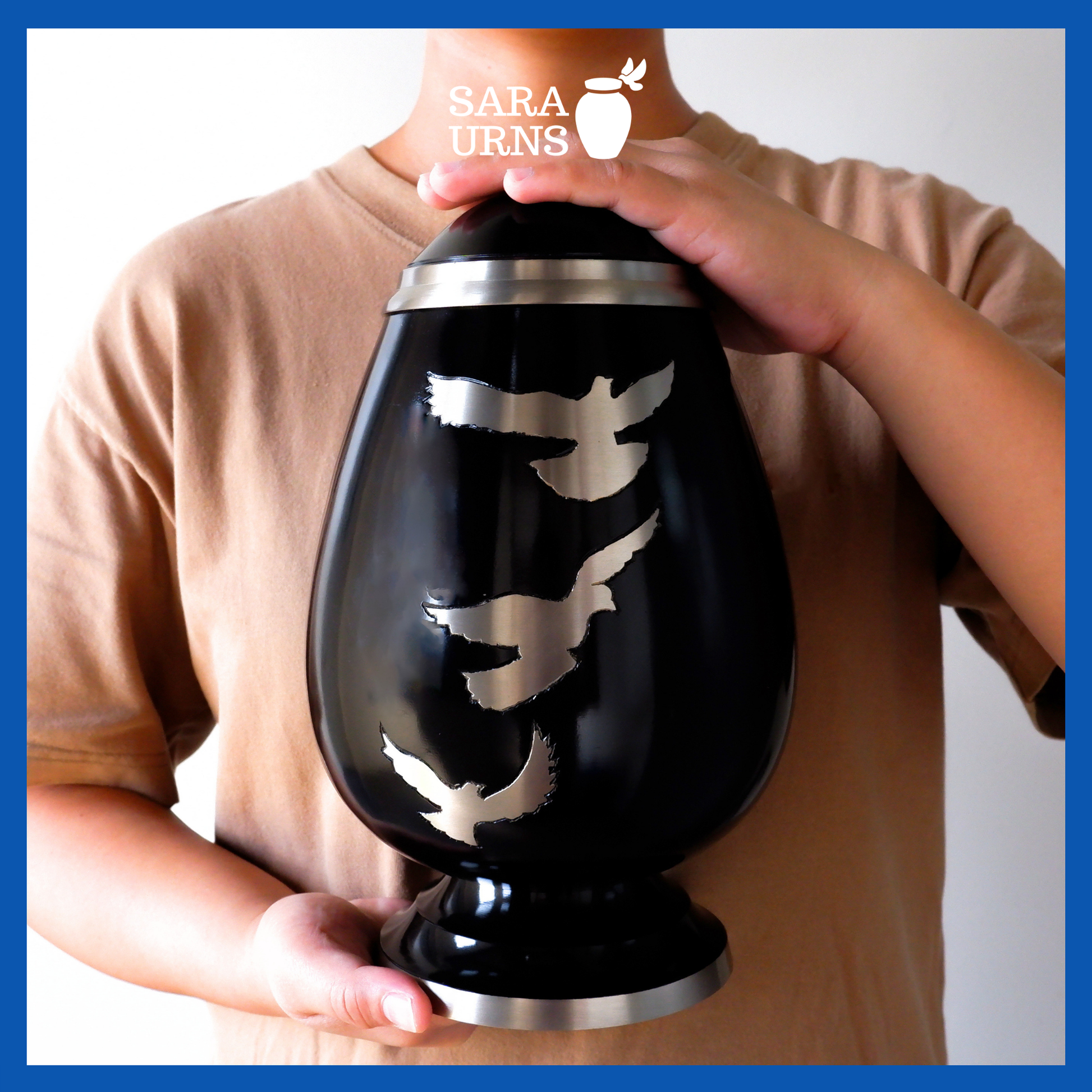 A presentation of a black polished brass metal urn with a silver, hand-etched detail of soaring doves held by an adult woman with two hands.