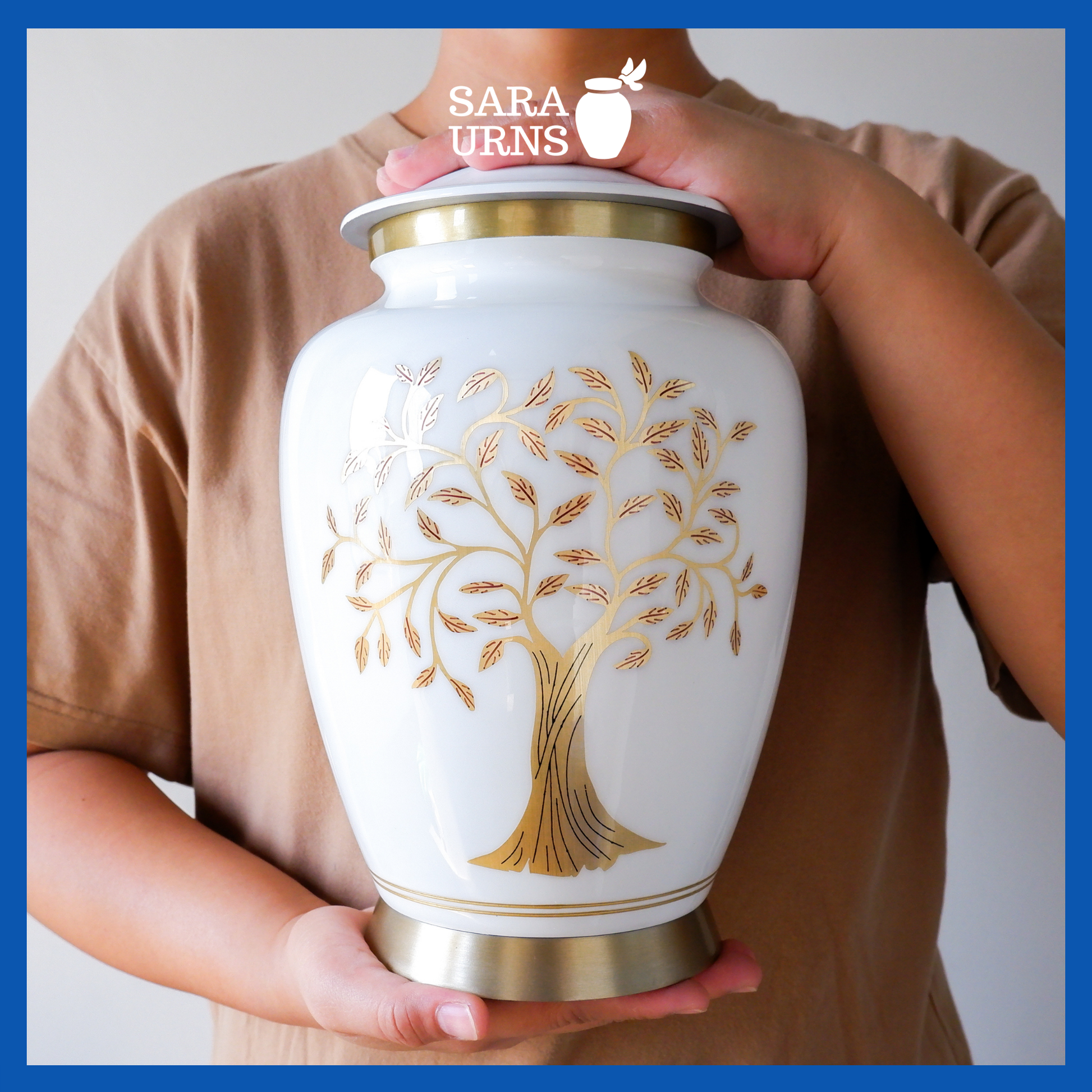 A presentation of a white polished aluminum metal urn with a golden, hand-etched tree of life detail held by an adult woman with two hands.